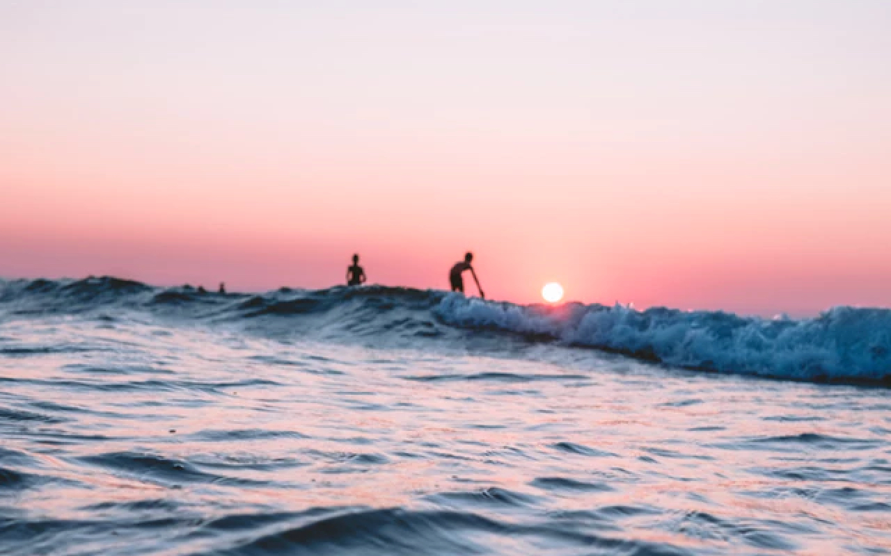 silhouettes of 2 guys surfing some gnarly waves at sunset against a pink sky